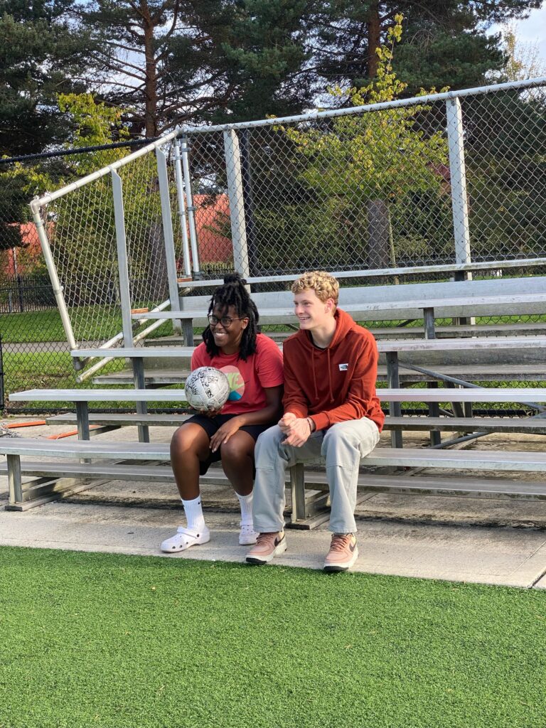 Senior photographer at Edmonds Woodway High School guiding a senior boy during his session, demonstrating posing techniques with a ball. Both photographer and senior are laughing together, showcasing their rapport and ensuring a stress-free senior photo experience. 